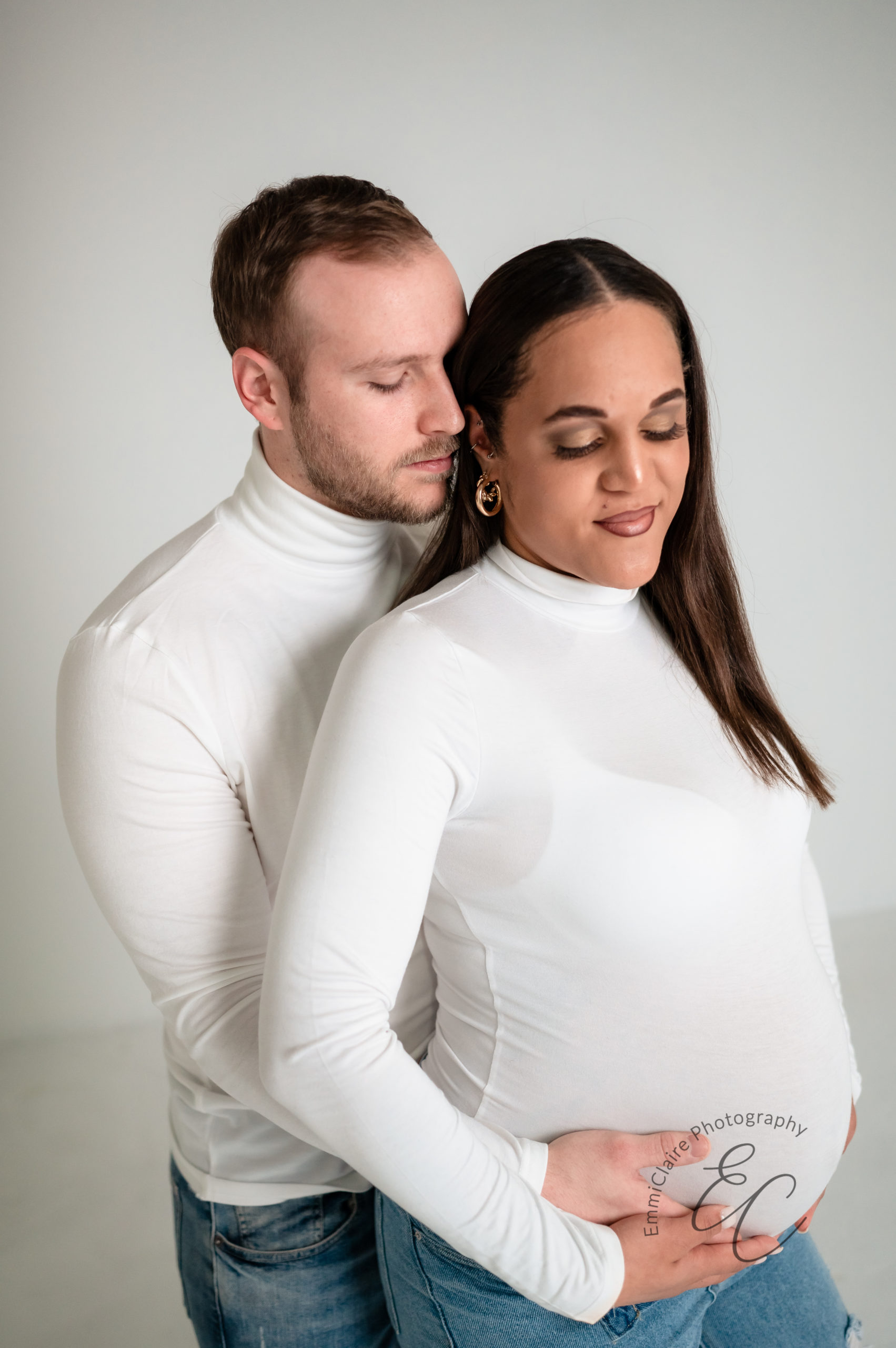 Pregnant woman, gently holding her swollen belly gazes down while her husband nuzzles in close to her ear, also holding her belly during their mellow studio maternity session in Richmond VA