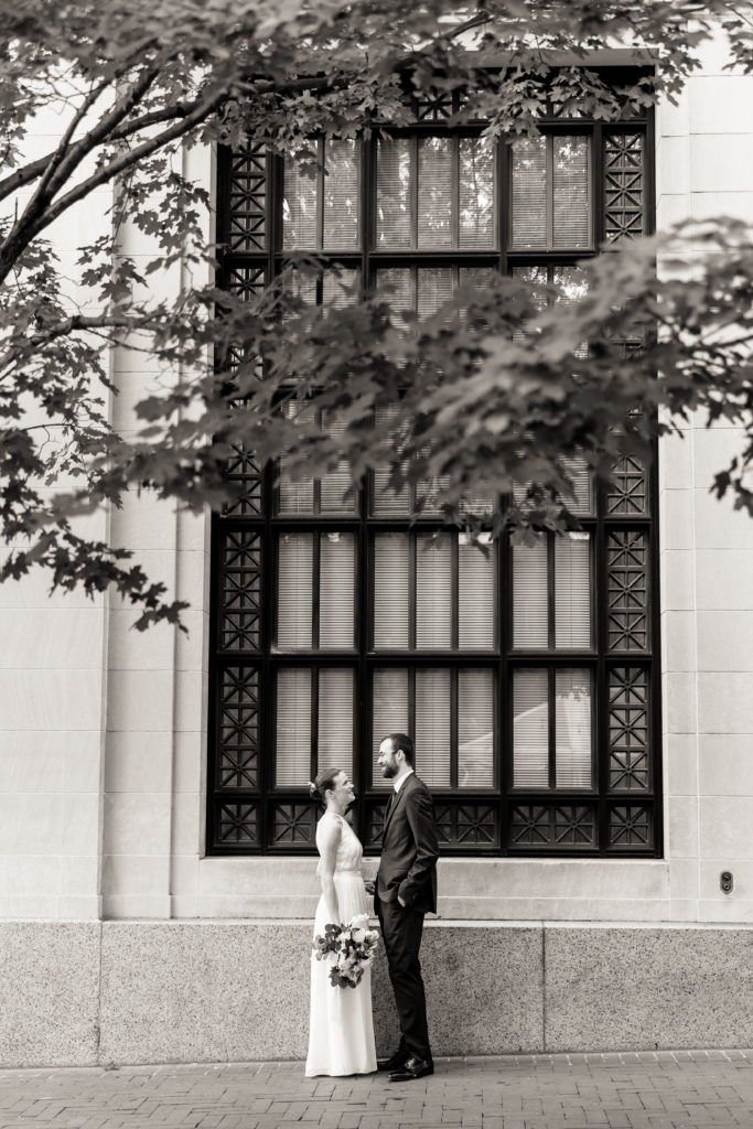 The couple stand in front of a large ornate window, gazing lovingly at one another immediately before their atrium wedding ceremony