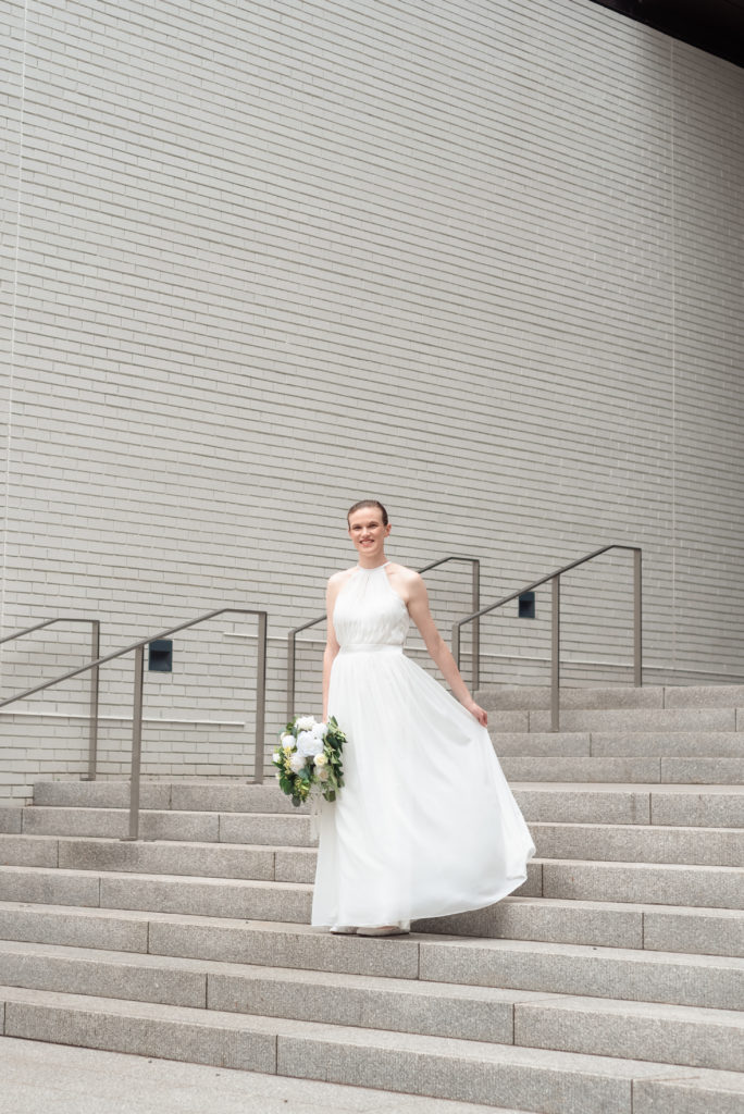 The bride gracefully ascends a set of modern stone steps, one hand caresses her gown as the other holds her bouquet.
