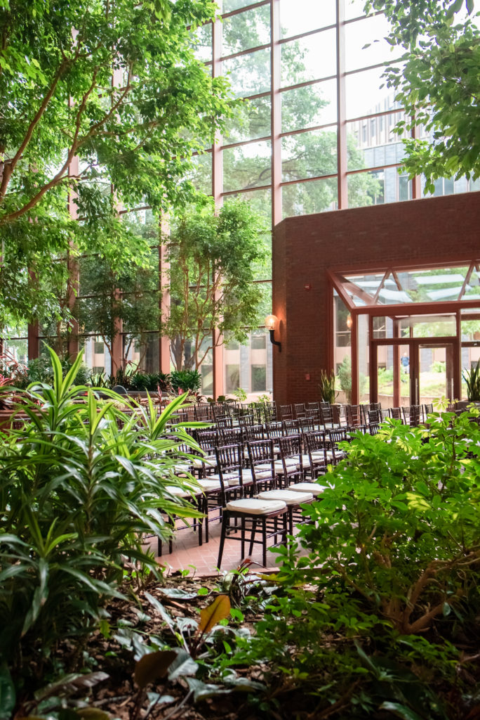Dark chiavari chairs, surrounded by lush greenery, trees, and shrubs, await guests for an atrium wedding ceremony.