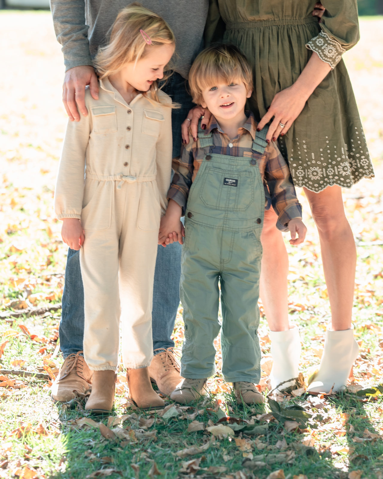 Mom and dad's hands rest on son and daughters shoulders during annual family portrait session. Sister gazes down at her brother, who is holding her hand while he gazes directly at the camera.