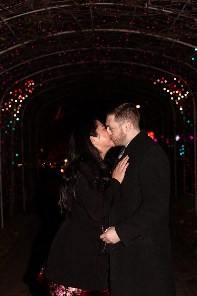 Terri and Kevin holding hands with the glowing holiday lights of Lewis Ginter GardenFest in the background, photographed by EmmiClaire Photography.