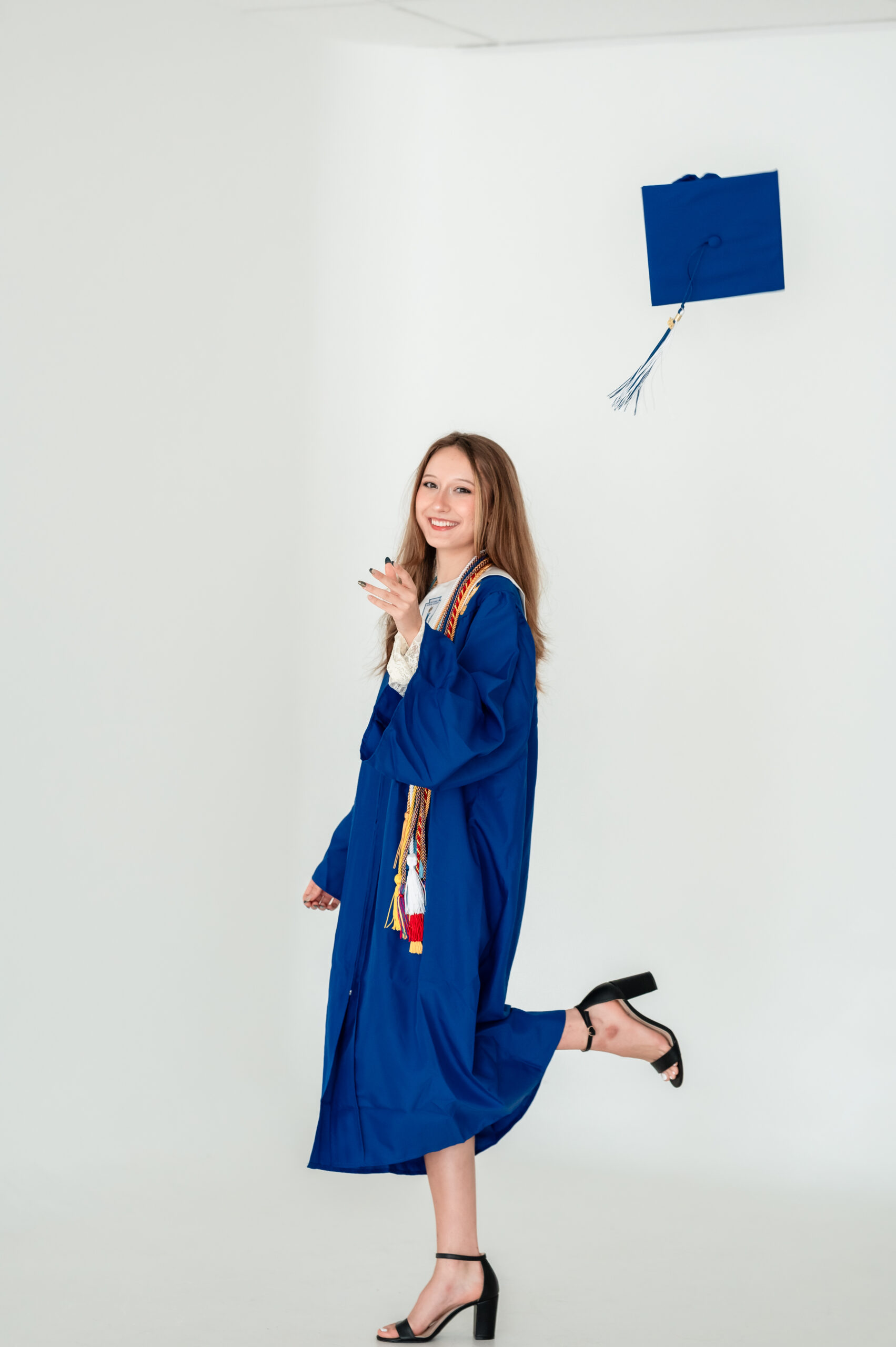 High school senior Anastasia smiles while tossing her graduation cap in a white studio, wearing a royal blue cap and gown with honor cords.