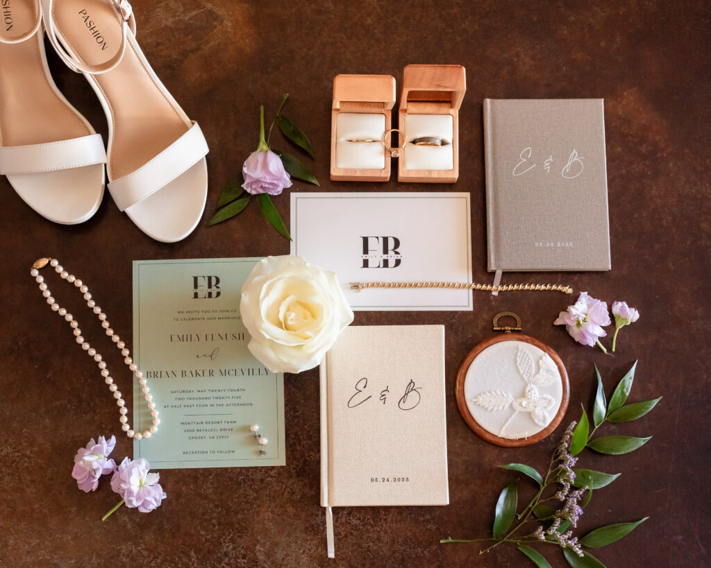 Flat lay of wedding day details featuring pearl jewelry, white sandals, gold rings in boxes, custom vow books, floral invitation suite, and soft lavender accents.