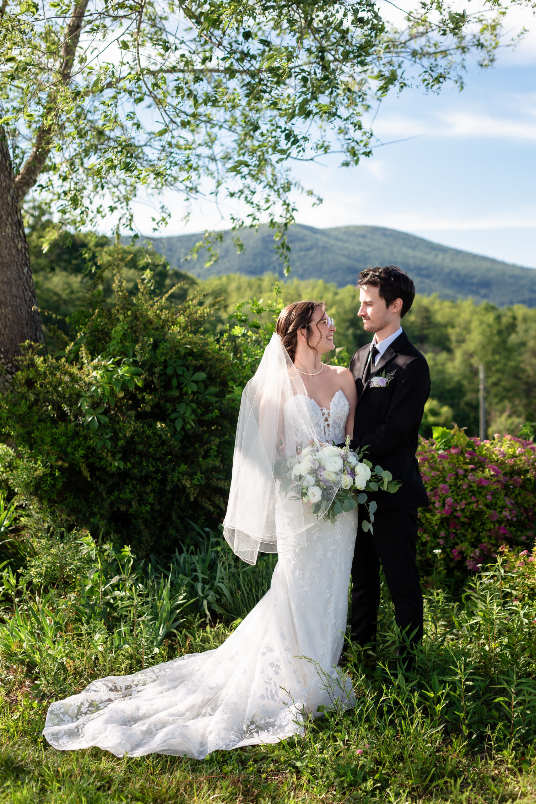 Bride and groom standing in front of the Blue Ridge Mountains at Montfair Resort Farm, smiling at each other under soft light and greenery.