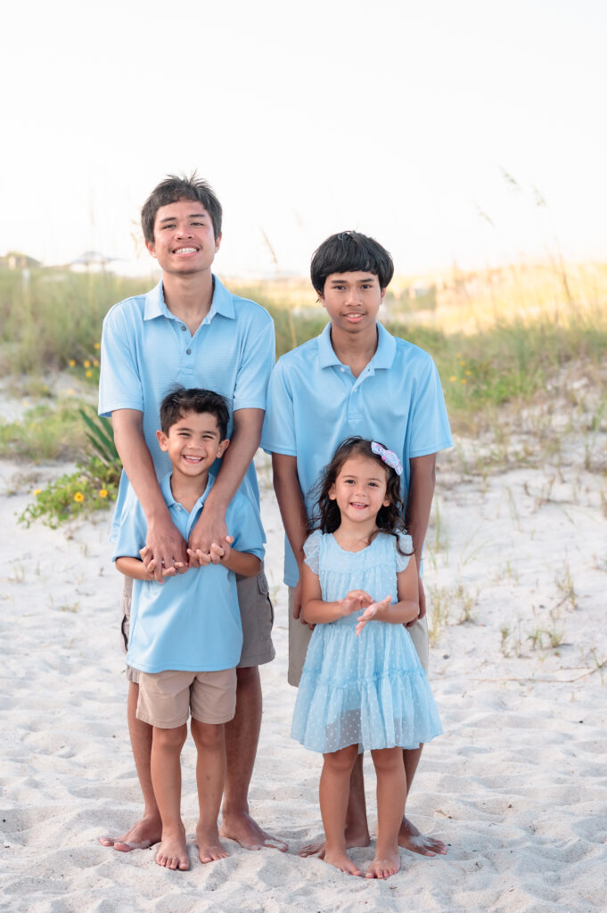 Four cousins in blue outfits standing close together on the sand, laughing and enjoying the beach.