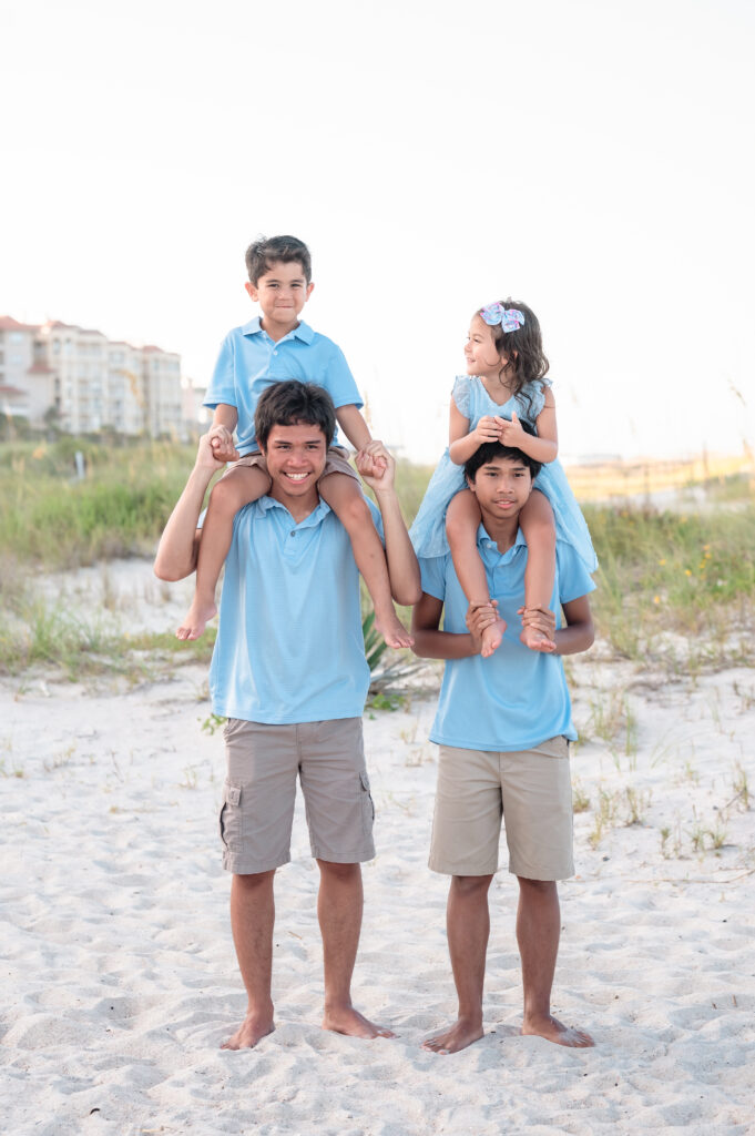 Four cousins in blue outfits sitting close together on the sand, laughing and enjoying the beach.