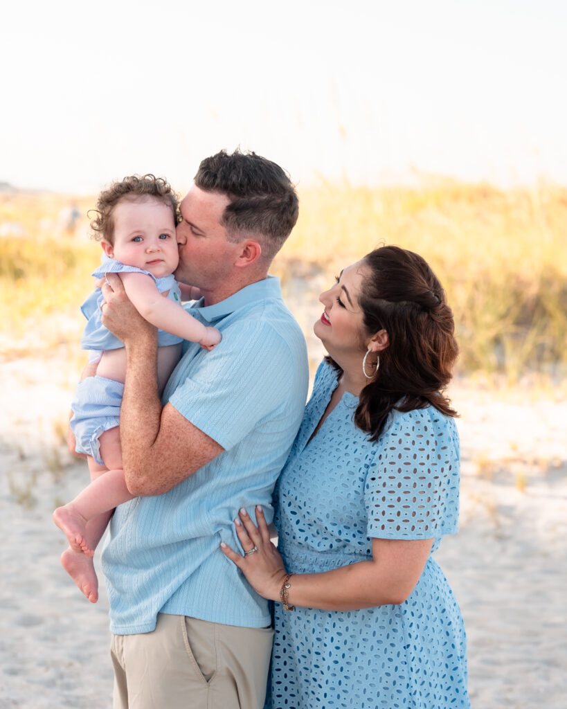 Parents holding their baby on the shoreline with waves and a pastel sunset sky behind them.