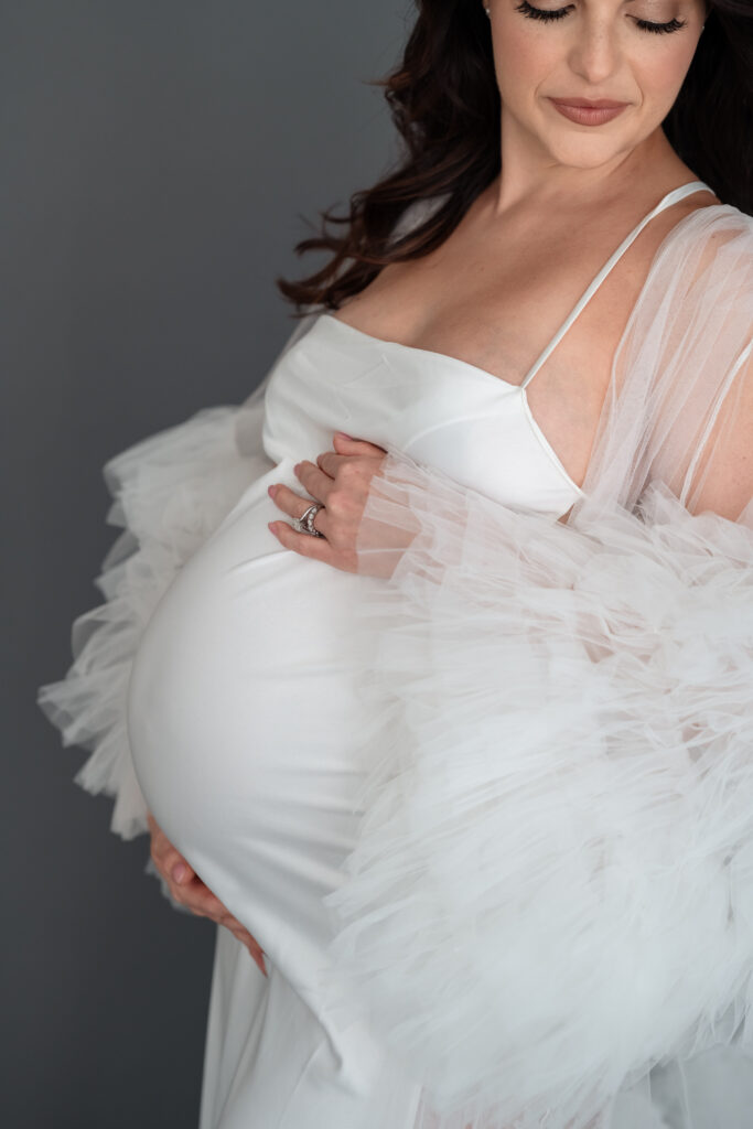 Mom posing in white gown and tulle robe during studio maternity session.