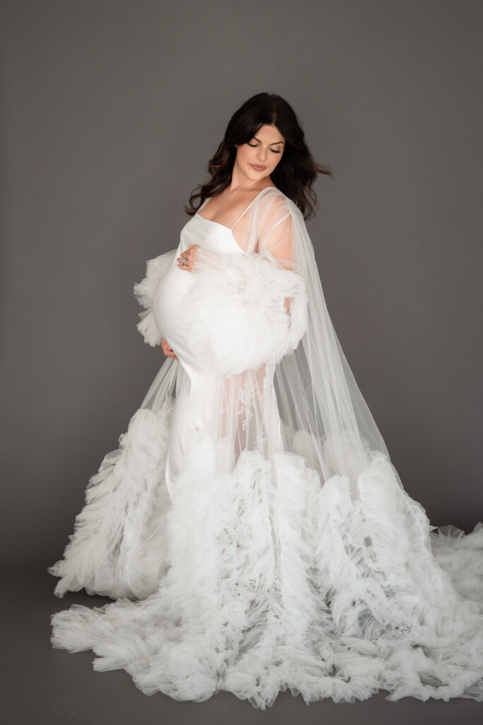 Mom posing in elegant white gown during studio maternity session.