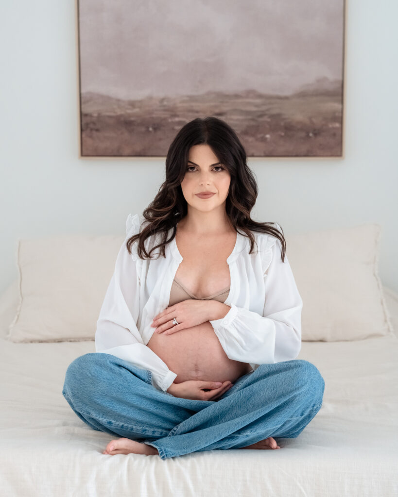 Mom sitting on cream bed touching hair during studio maternity session.