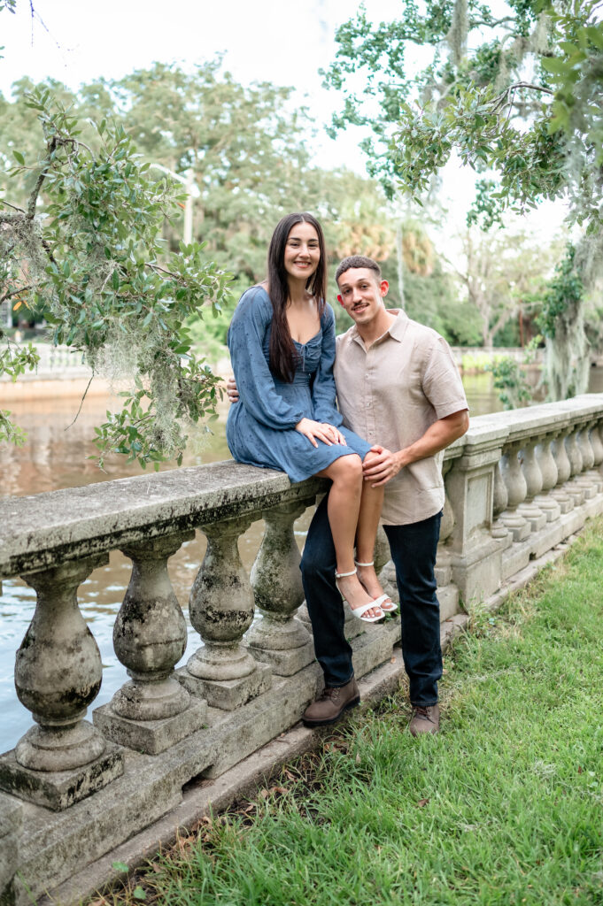 Newly engaged couple sitting along stone railing overlooking the water