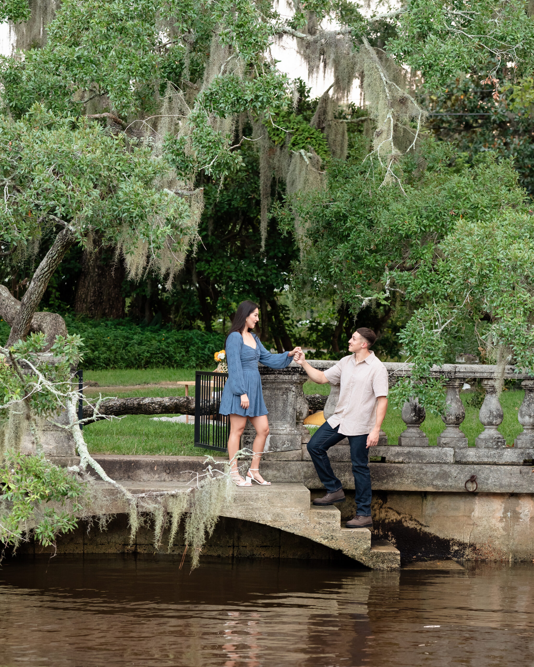 Engaged couple sitting by the water at Yacht Basin Park with stone railing and Spanish moss trees in background.